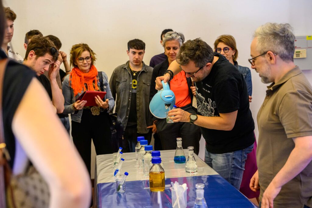 in un laboratorio di scienza per il pubblico, un divulgatore versa acqua calda per un esperimento, mentre il pubblico lo guarda