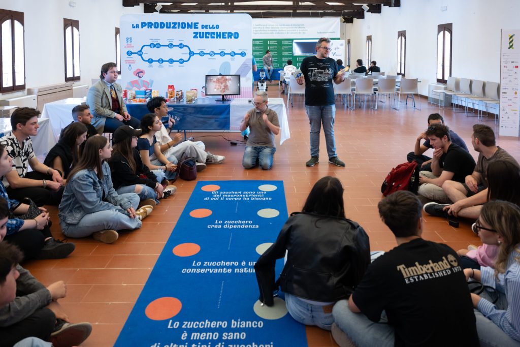 in un laboratorio di scienza per il pubblico, una scolaresca ascolta i due divulgatori scientifici che parlano
